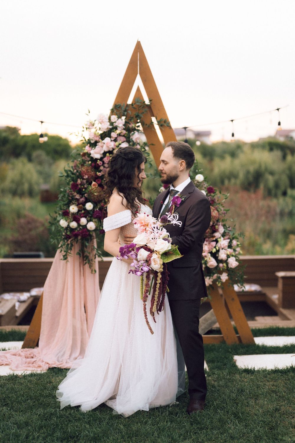 Couple exchanges vows at outdoor wedding ceremony beneath triangular wooden arch adorned with pink and burgundy floral arrangements.