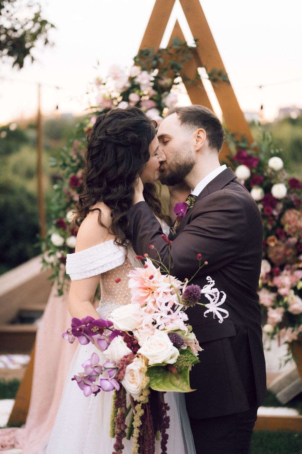 Bride and groom sharing a kiss at outdoor wedding ceremony with floral arch backdrop and bridal bouquet.
