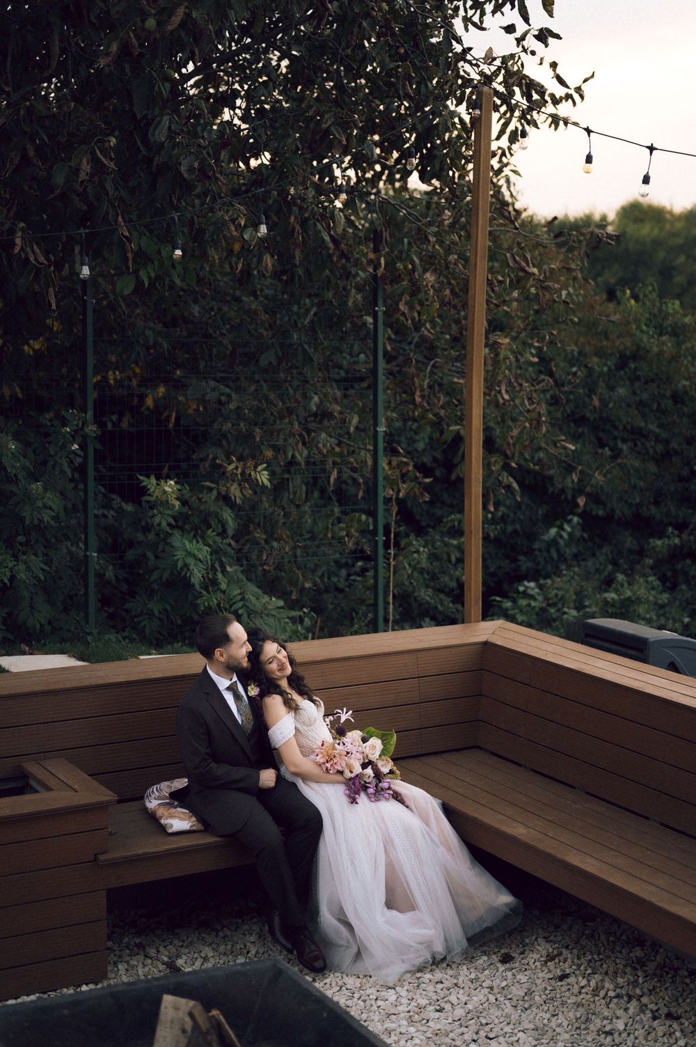 Bride and groom sit closely on a wooden bench surrounded by lush greenery at dusk, bride holding a purple bouquet.