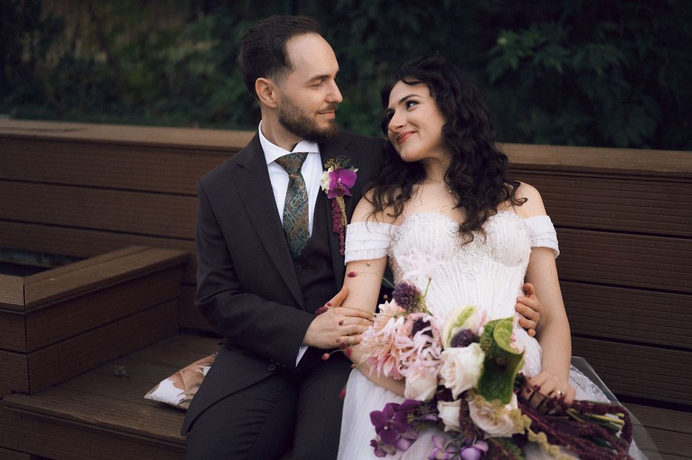 A bride and groom sit together on a wooden bench, smiling at each other, as she holds a purple and pink floral bouquet.