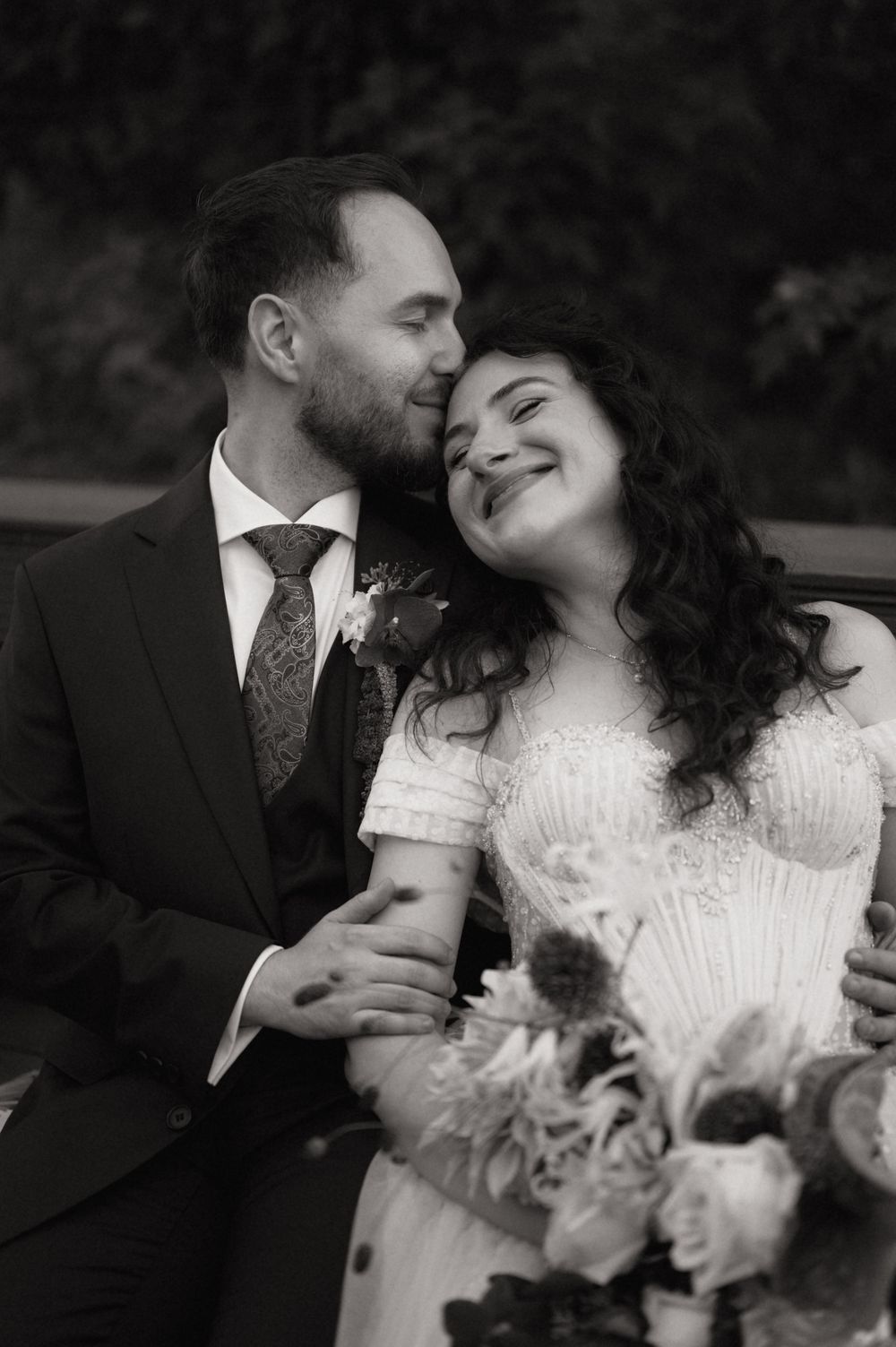 Black and white wedding photo of a smiling bride holding bouquet as groom kisses her cheek tenderly.