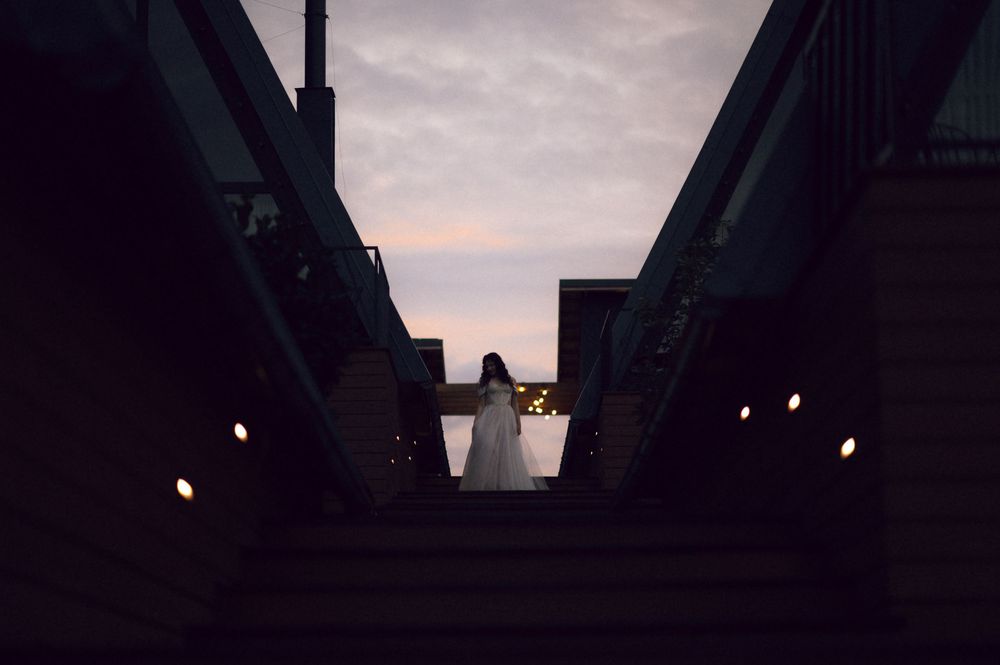 Woman in white dress standing on stairs between two bridges at dusk with glowing lights.