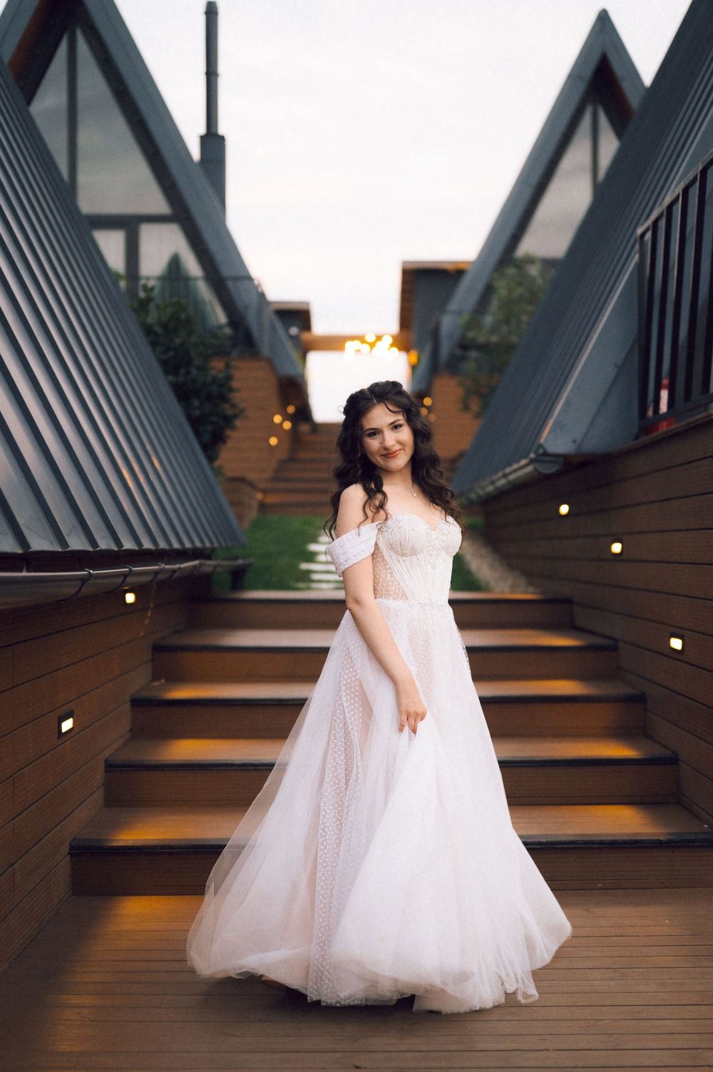 A bride in an off-shoulder white tulle gown poses gracefully on wooden stairs at dusk.