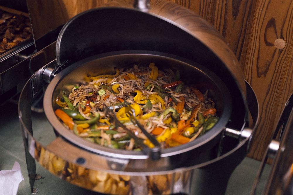 Colorful stir-fry vegetables in a buffet chafing dish with lid open, featuring peppers and onions.