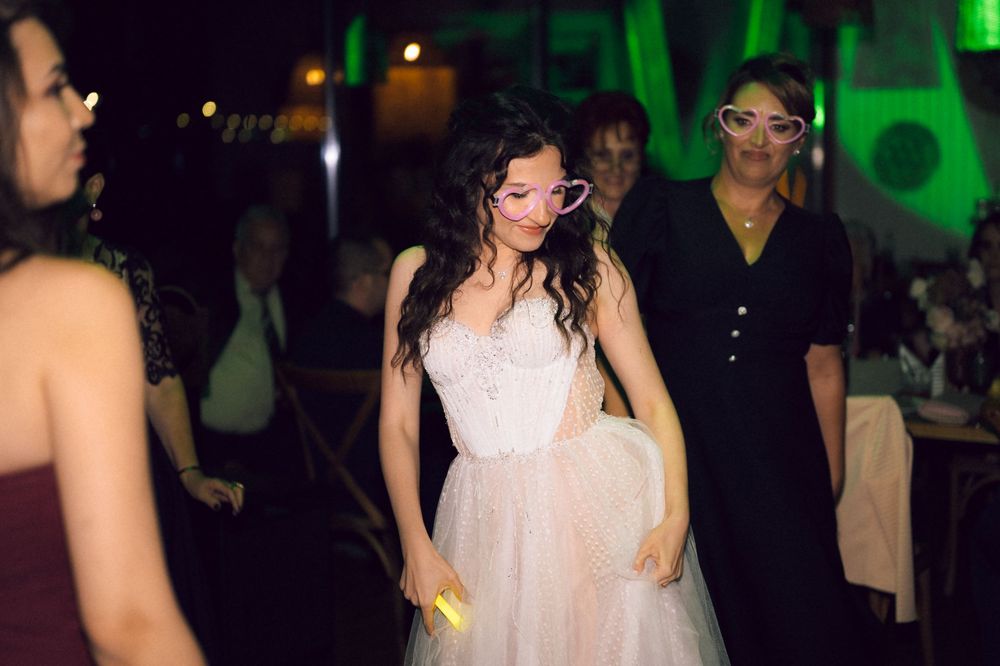 Woman in pink tulle dress dancing at wedding reception with guests wearing novelty glasses under green lights.