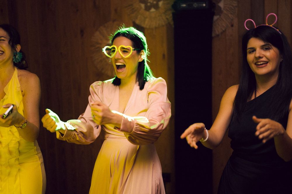 Three women dancing and laughing at a party, wearing colorful outfits including heart-shaped sunglasses and bunny ears.