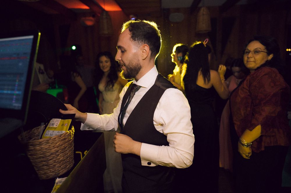 A bearded DJ in a vest manages a digital music setup at a lively indoor event with guests in the background.