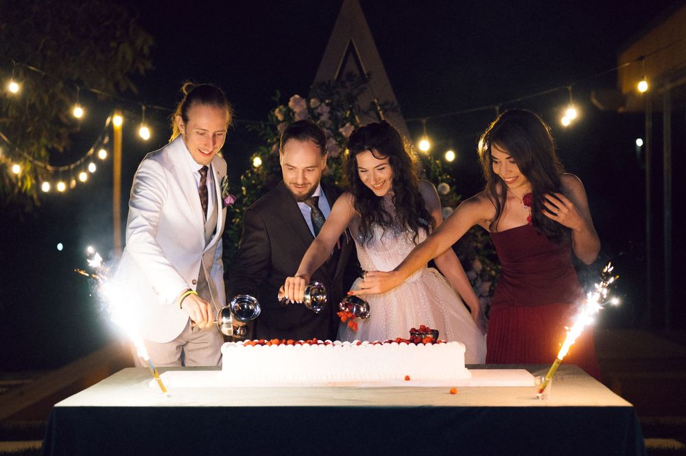 Four people cutting a white wedding cake at night with sparklers and string lights in the background.