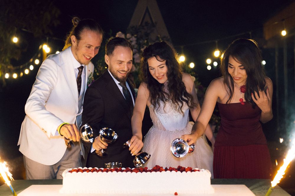 Four elegantly dressed guests cutting a white wedding cake outdoors at night with sparklers and string lights.