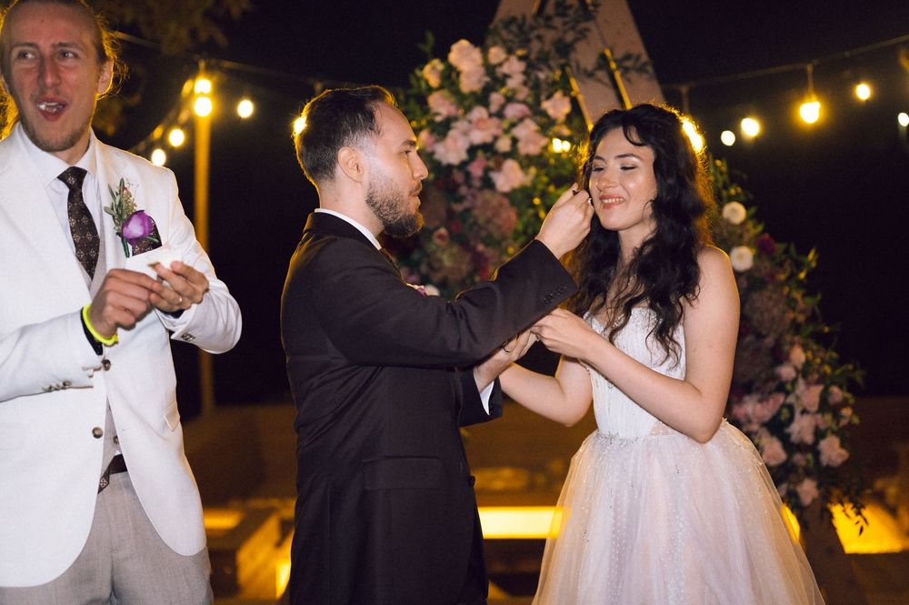 Groom placing wedding ring on brides finger during outdoor evening ceremony with string lights and floral arch.