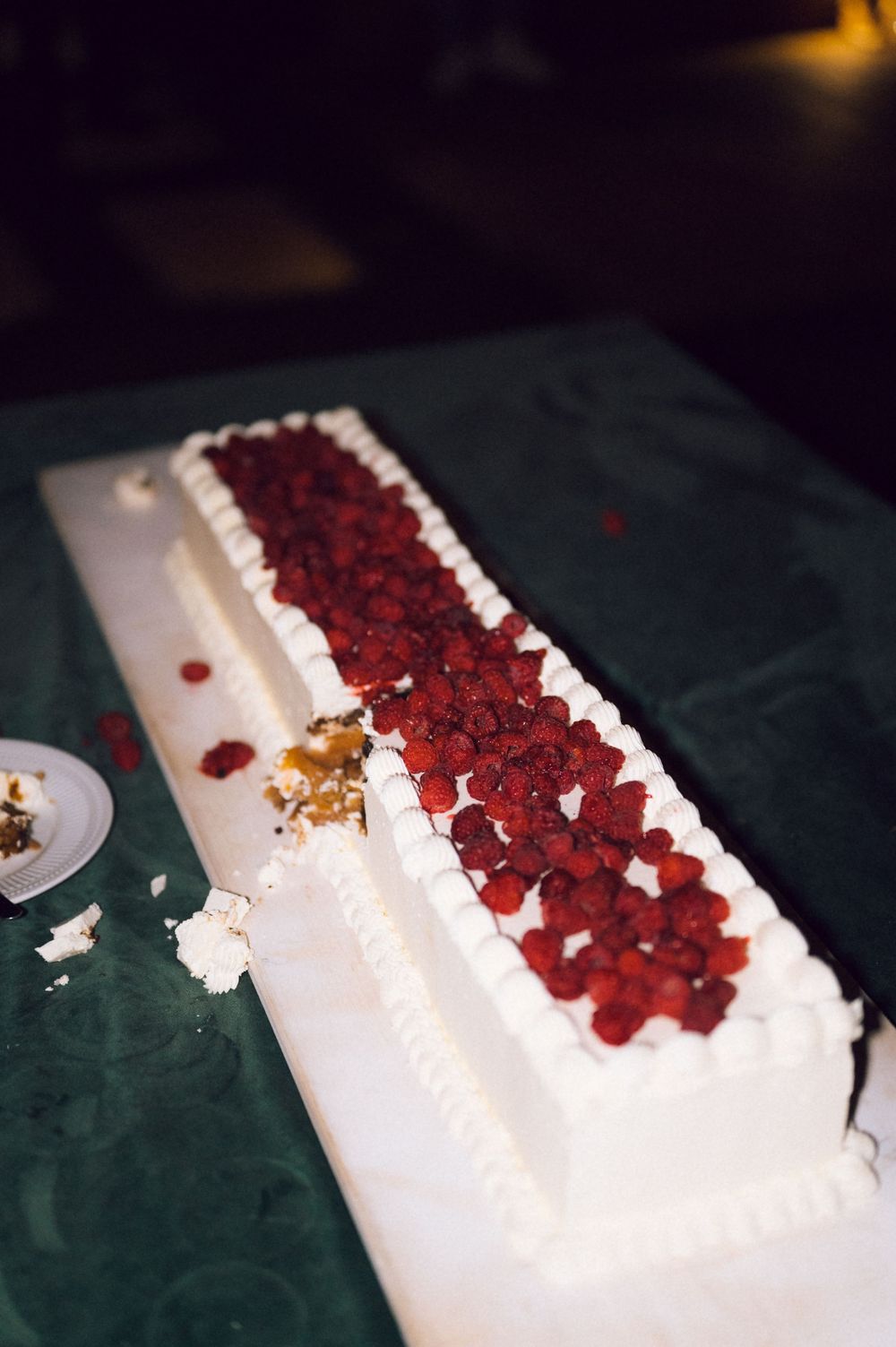 Elegant rectangular white wedding cake topped with fresh raspberries on a green tablecloth, partially cut.
