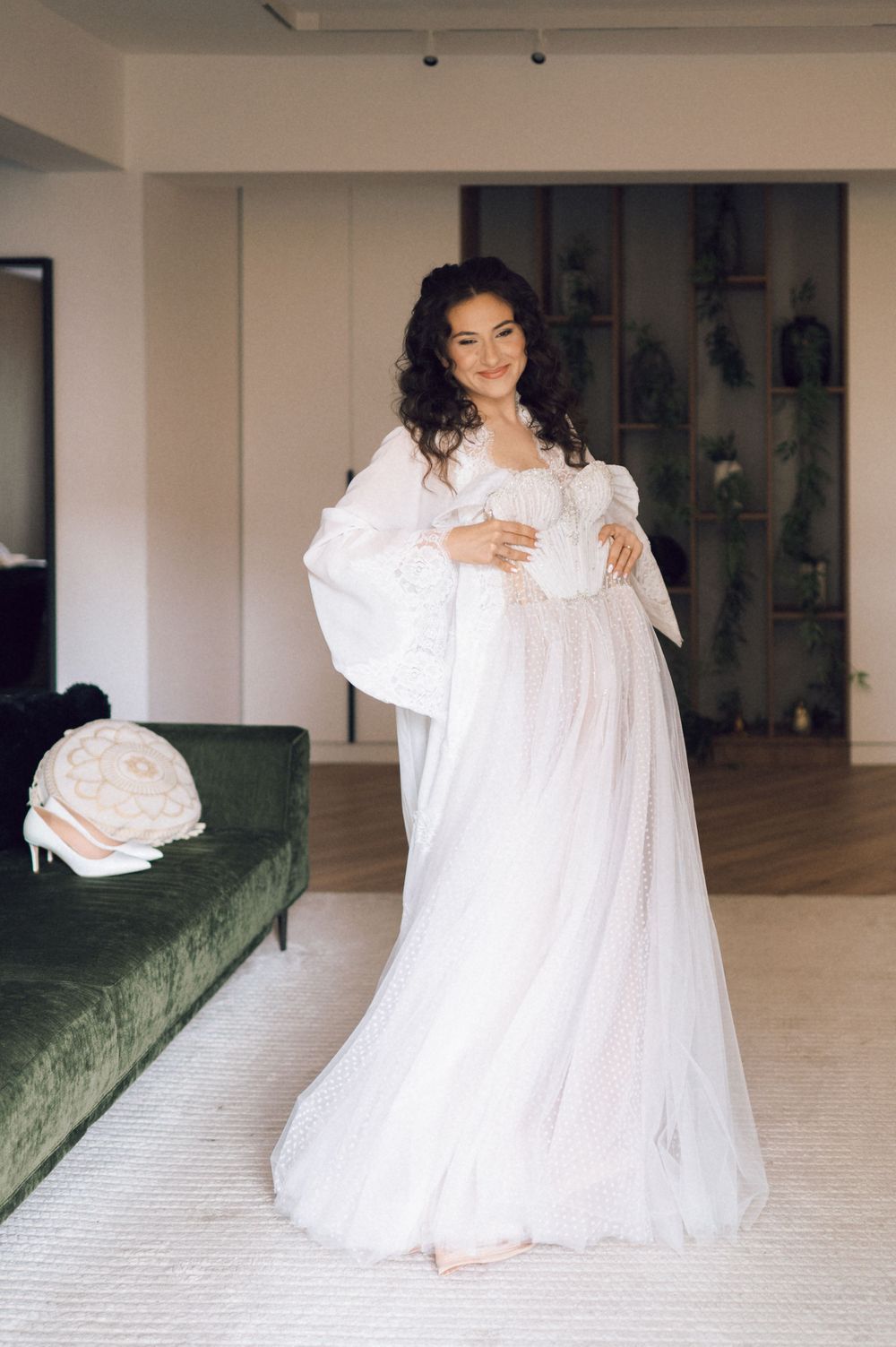 Bride in elegant white tulle gown with sheer sleeves posing indoors near green velvet sofa.