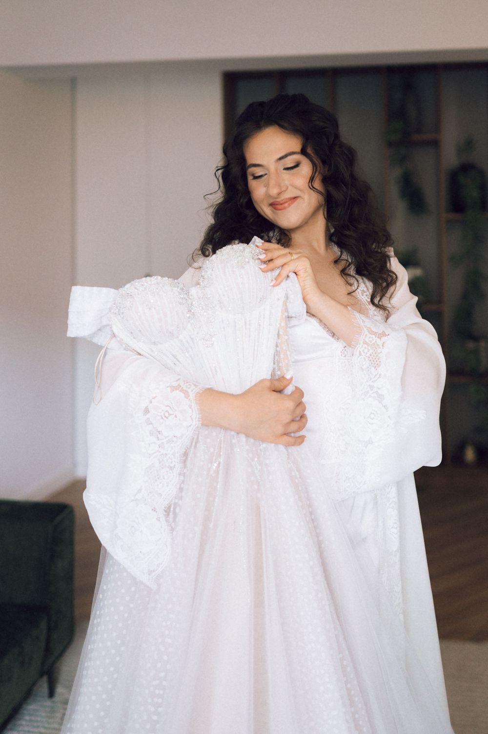 Smiling bride in elegant white lace wedding gown with sheer sleeves, holding her dress in a bright indoor setting.