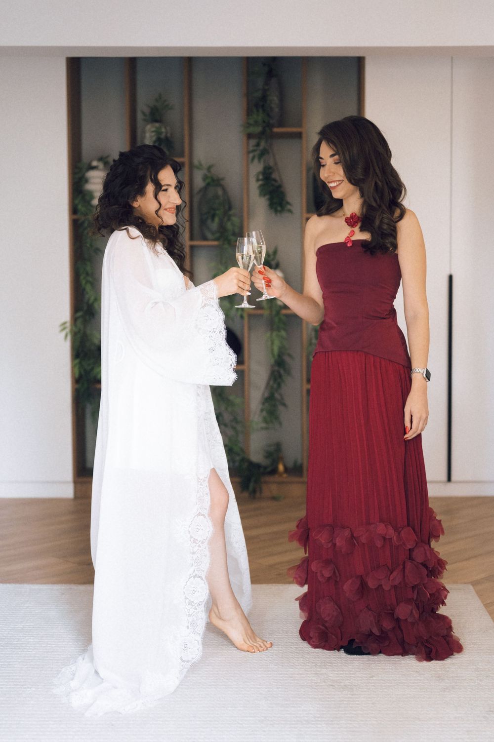 Two women toasting with champagne glasses, one in a white bridal robe and one in a burgundy gown, indoors.