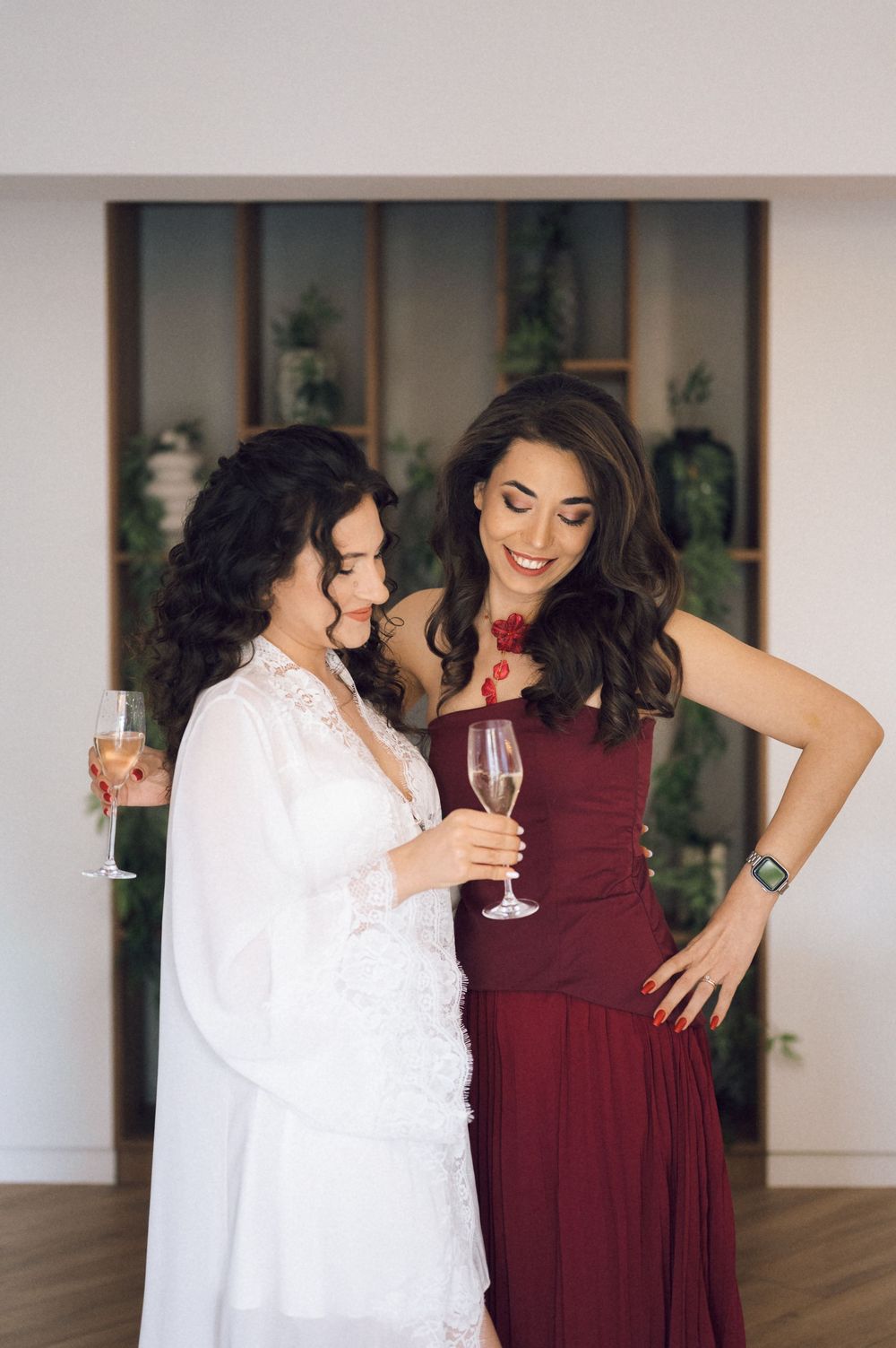 Two elegant women in formal attire toasting with champagne glasses, one in a white bridal gown and one in a burgundy dress.