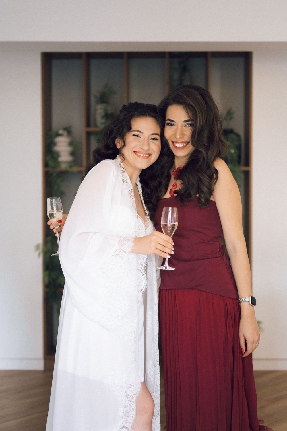 Bride in white lace gown and bridesmaid in burgundy dress toasting with champagne glasses at wedding reception.