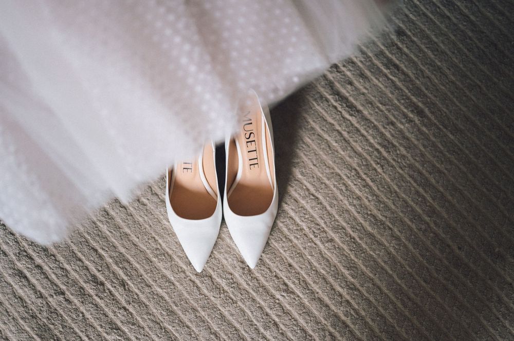 White pointed-toe bridal heels with gold insoles rest on a textured carpet beside a flowing white tulle wedding veil.