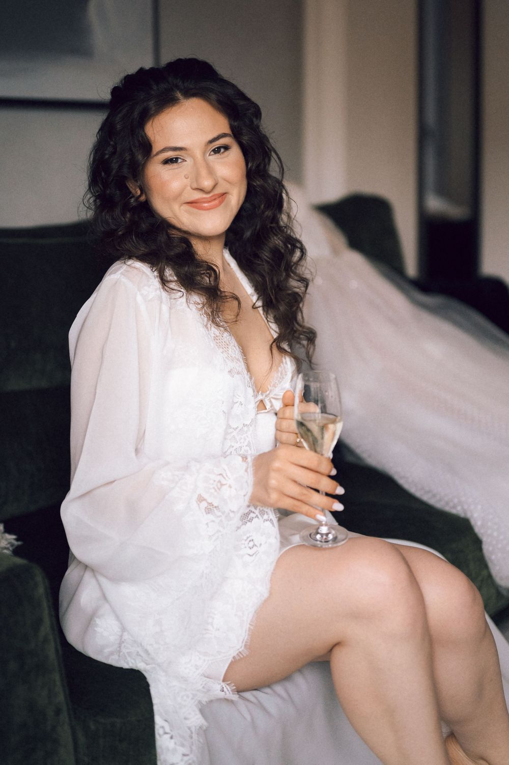 Smiling bride in white lace robe and veil sits on green sofa holding champagne glass.