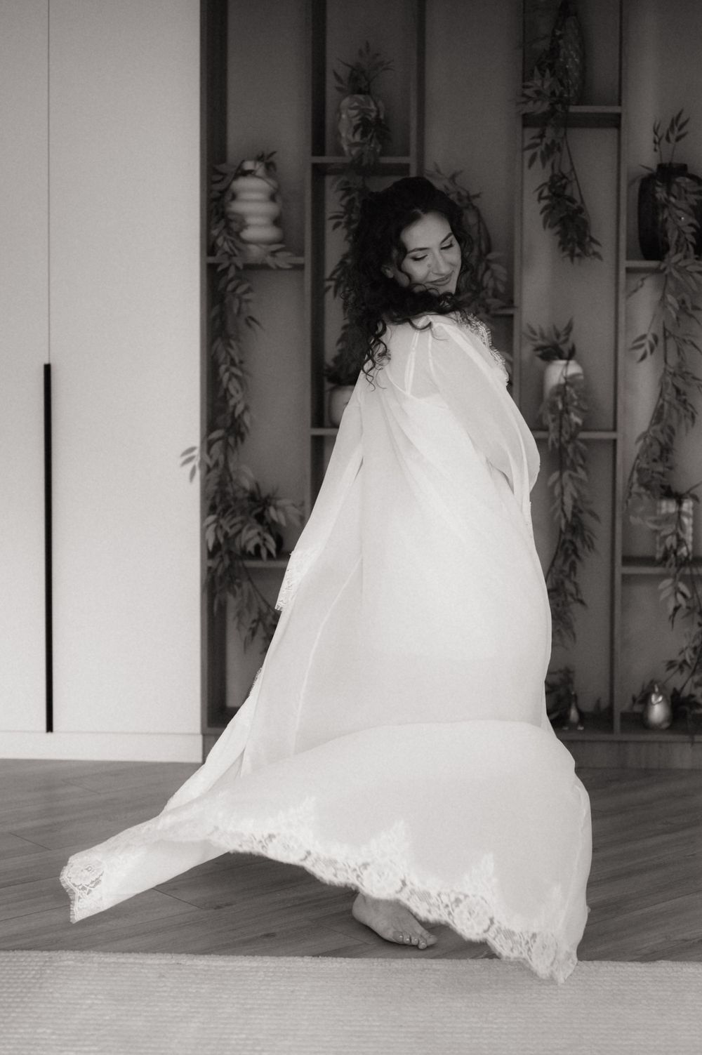Black and white photo of a woman in a flowing white lace gown twirling gracefully indoors near shelves with plants.