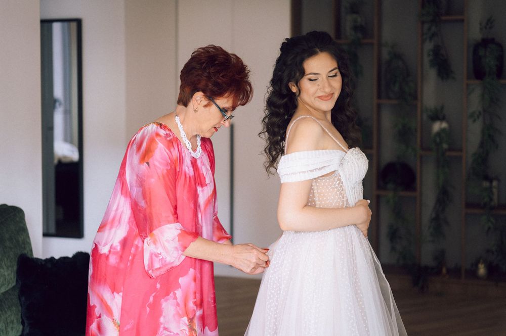 Older woman in pink floral dress helping a bride in white off-shoulder gown get dressed before wedding ceremony.