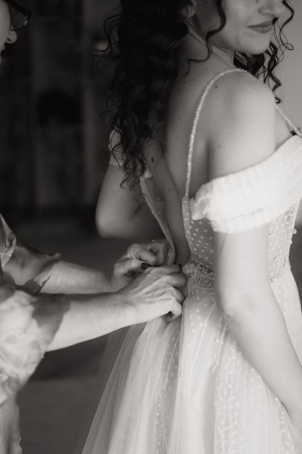 Black and white photo of a bride getting her wedding dress buttoned up by a helper before the ceremony.