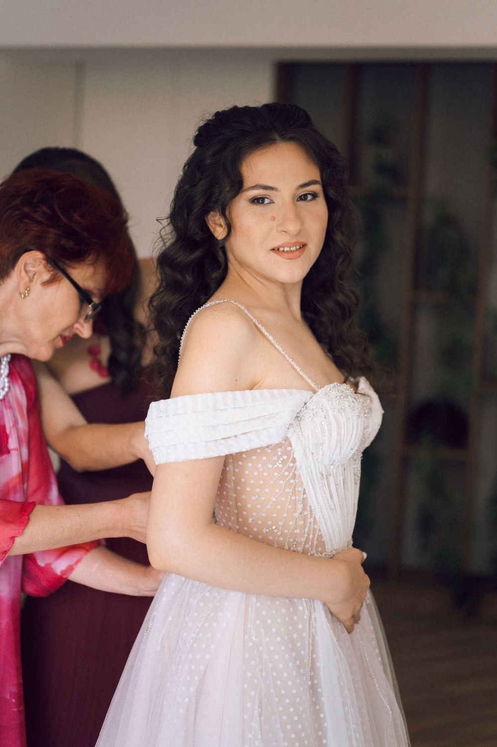 Bride in white off-shoulder polka dot tulle gown being helped by older woman during wedding preparations.