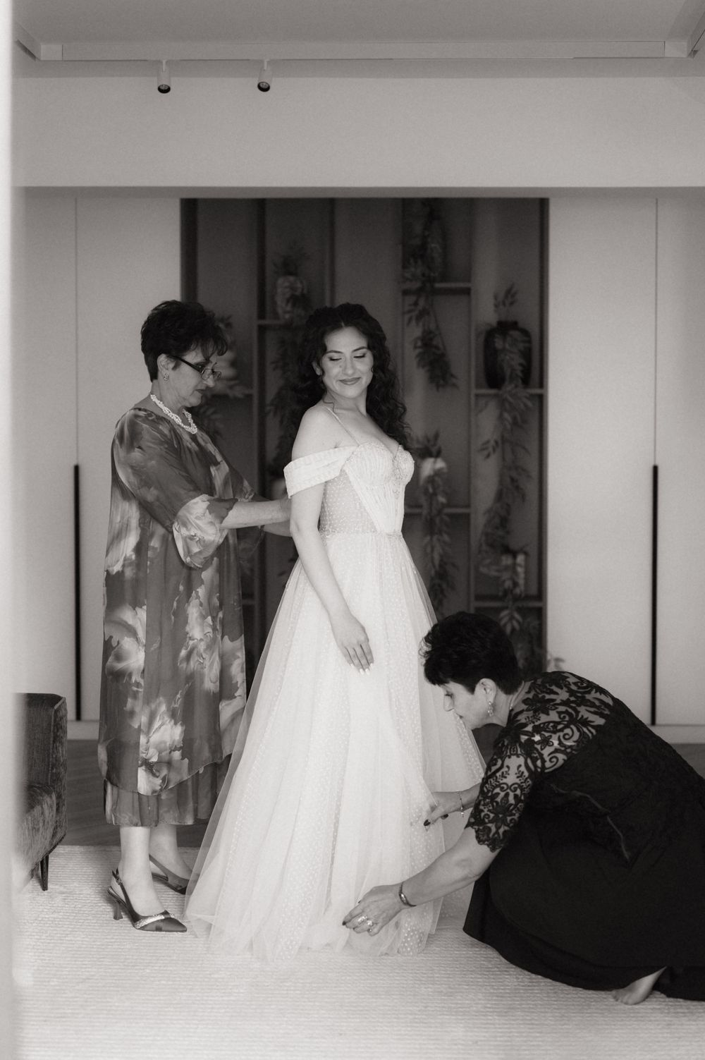 Black and white photo of a bride in an off-shoulder gown being helped by two people while getting ready.