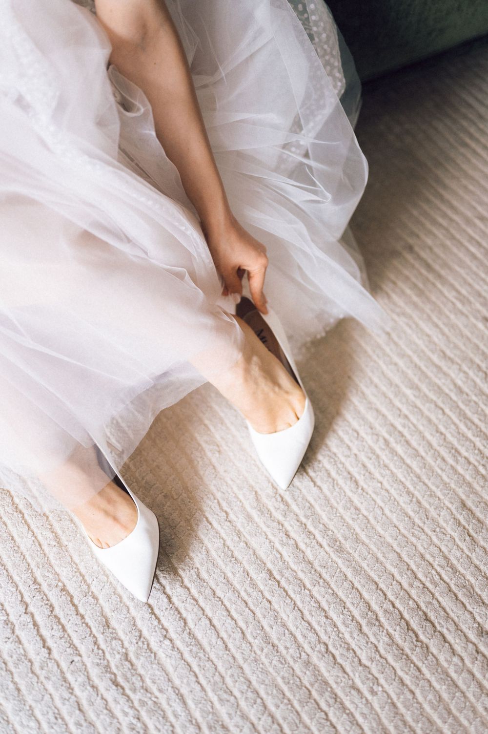 Bride in white tulle dress putting on elegant pointed-toe white heels on a cream carpet.
