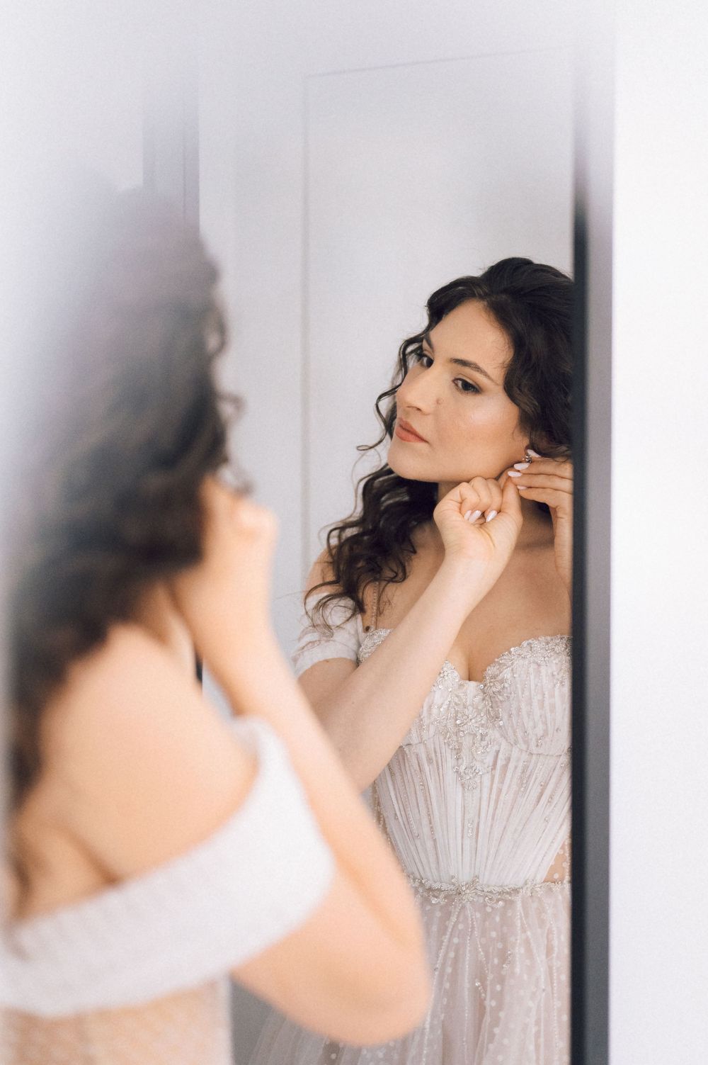 Bride in a beaded strapless wedding gown putting on earrings while gazing into a mirror.