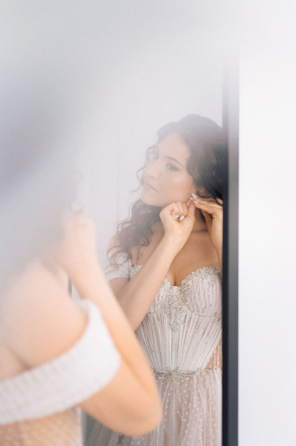 Bride in beaded strapless gown putting on earrings while looking in mirror before wedding ceremony.