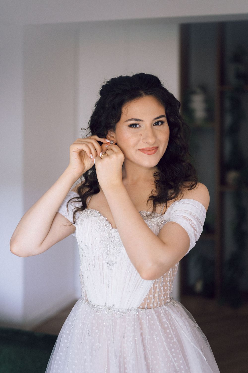 A smiling bride in a white off-shoulder beaded gown adjusts her earring while posing indoors.