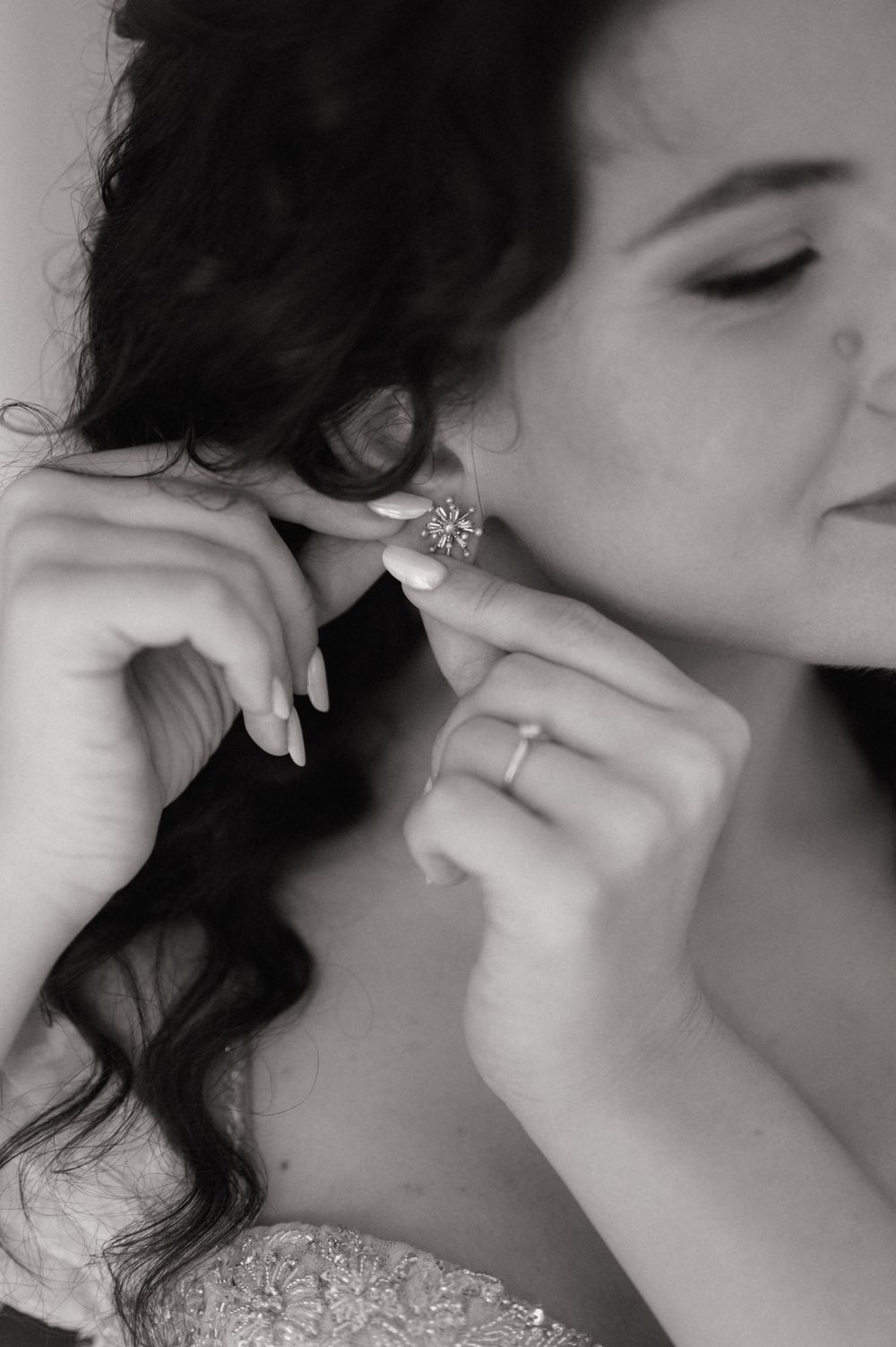 Bride in black and white photo putting on a sparkling earring, wearing an engagement ring and lace wedding dress.