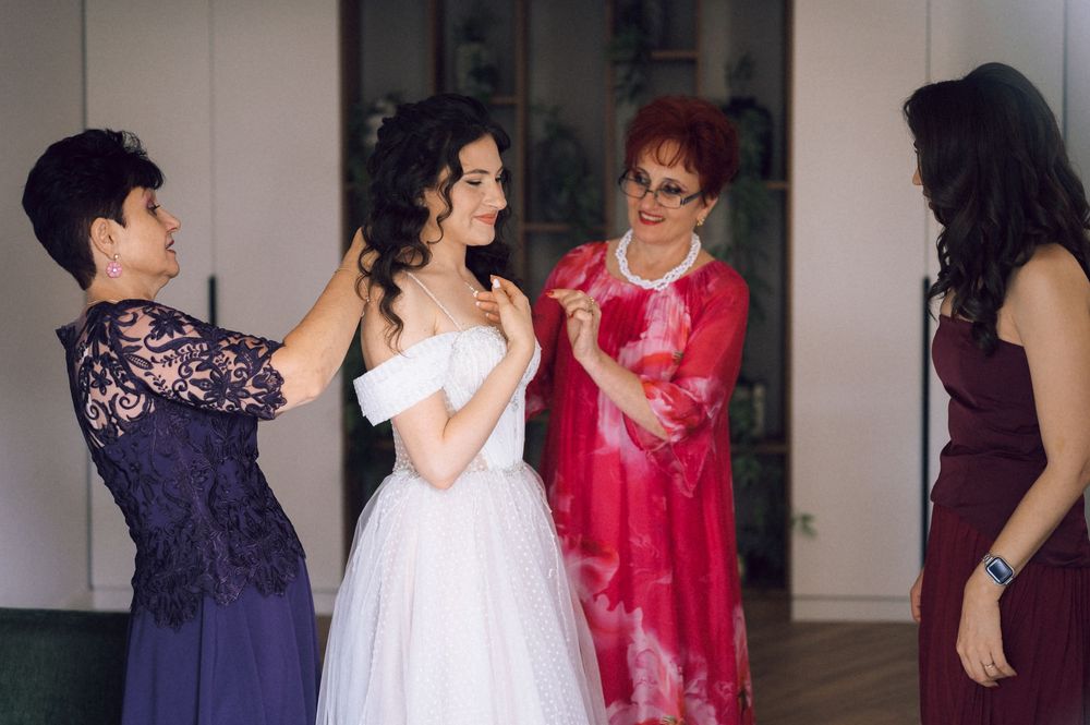 Bride in white gown surrounded by three women in formal dresses during wedding preparations.