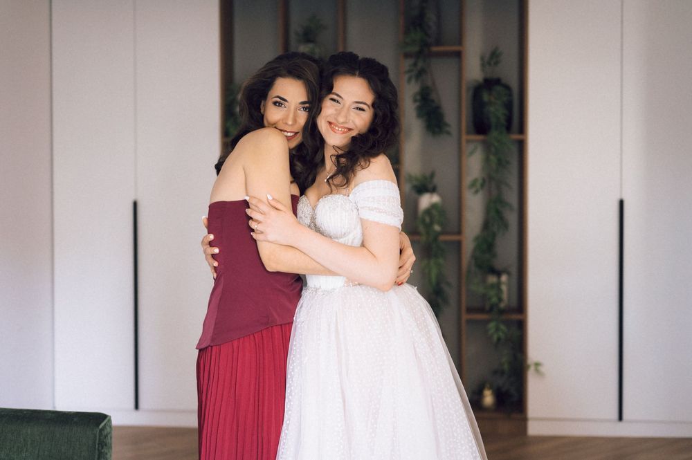 Two women embrace smiling, one in a white wedding gown and one in a red dress, posing indoors.
