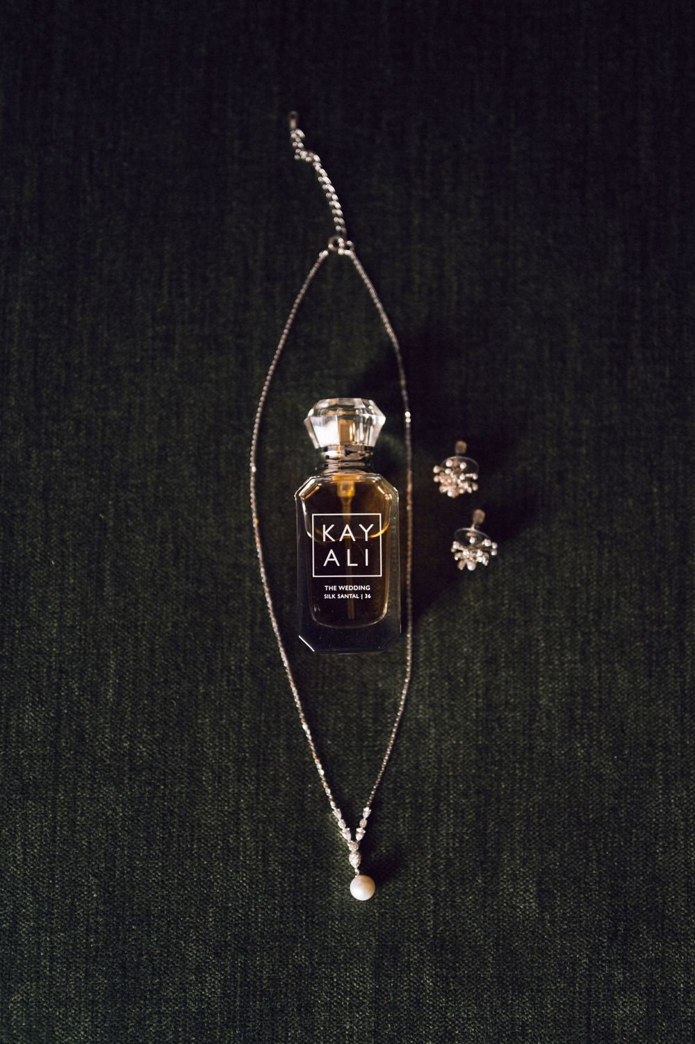 Kayali perfume bottle with diamond necklace and stud earrings on dark green fabric background.