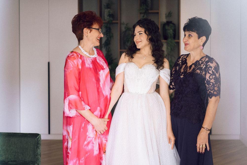 Bride in white off-shoulder gown stands between two women in formal attire, sharing a joyful moment before the wedding.