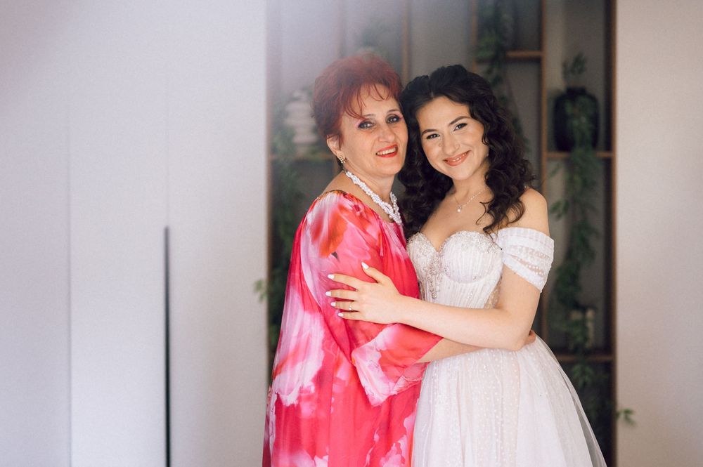 A bride in a white gown embraces an older woman in a pink floral wrap, posing together indoors.