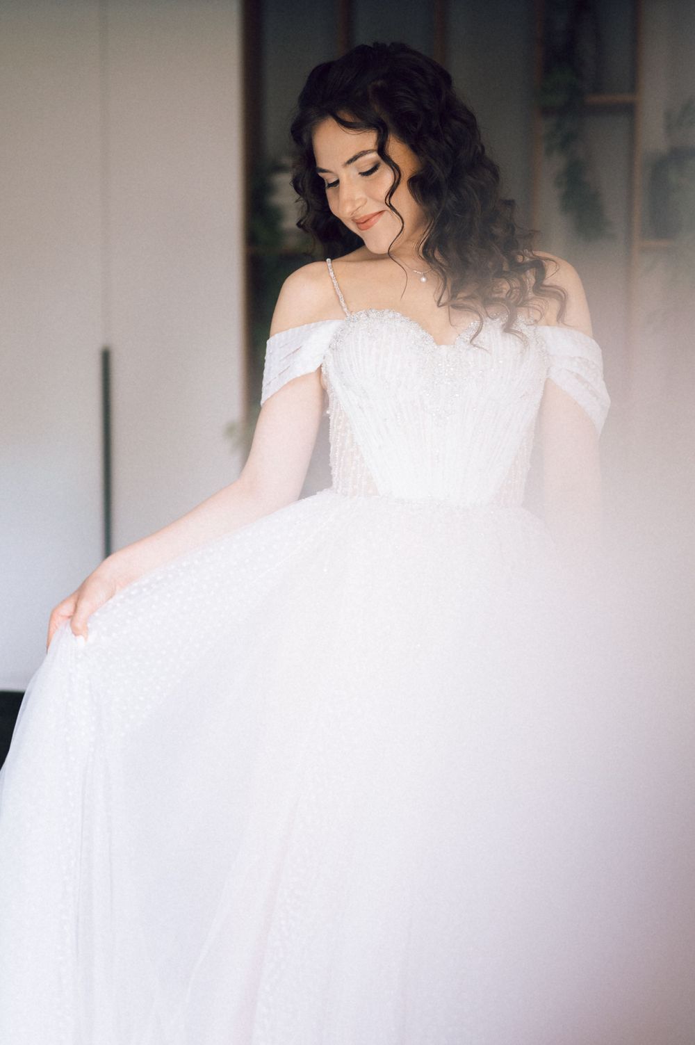 A bride in a white off-shoulder ball gown with lace bodice, holding her skirt, dark curly hair, soft natural light.