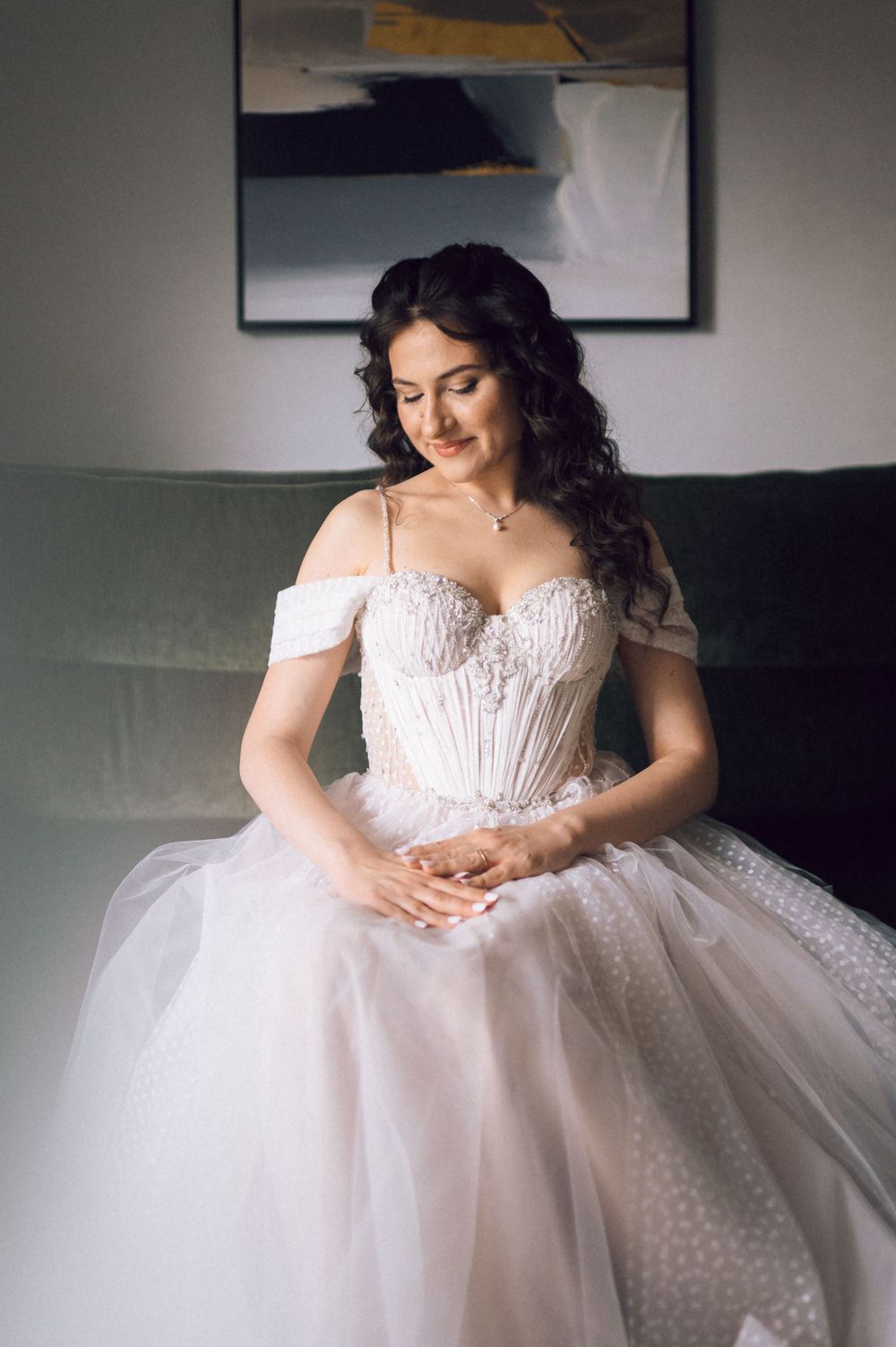 Bride in off-shoulder white lace ballgown sits gracefully on green sofa, smiling downward with elegant curled hair.