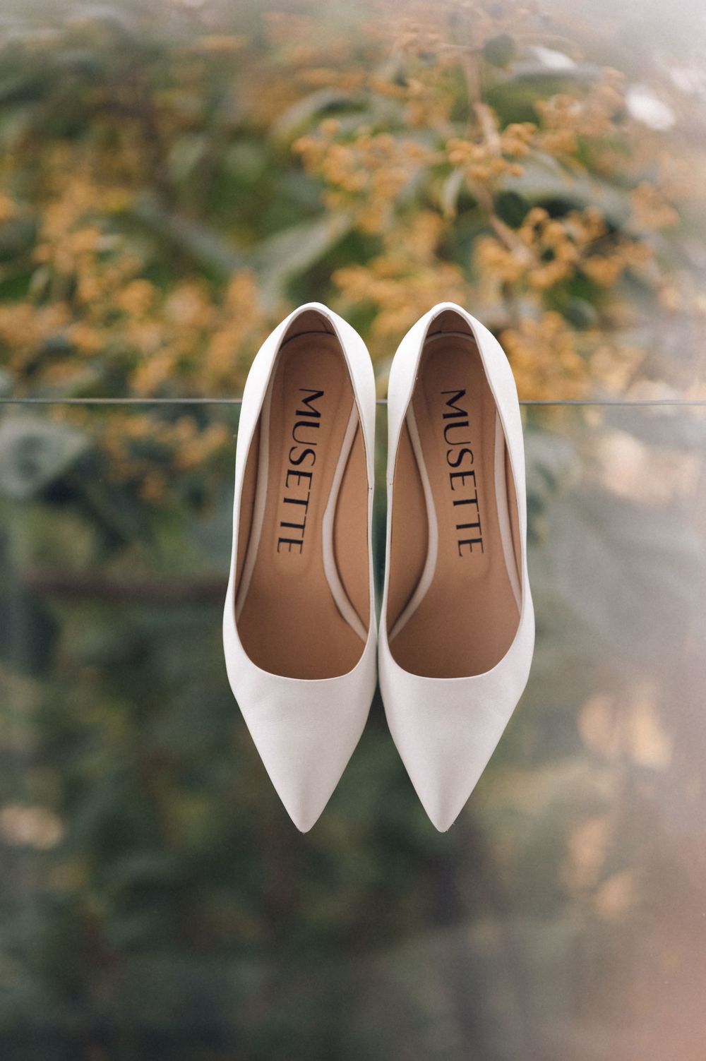 White Musette pointed-toe heels displayed on glass surface with blurred green and yellow foliage background.
