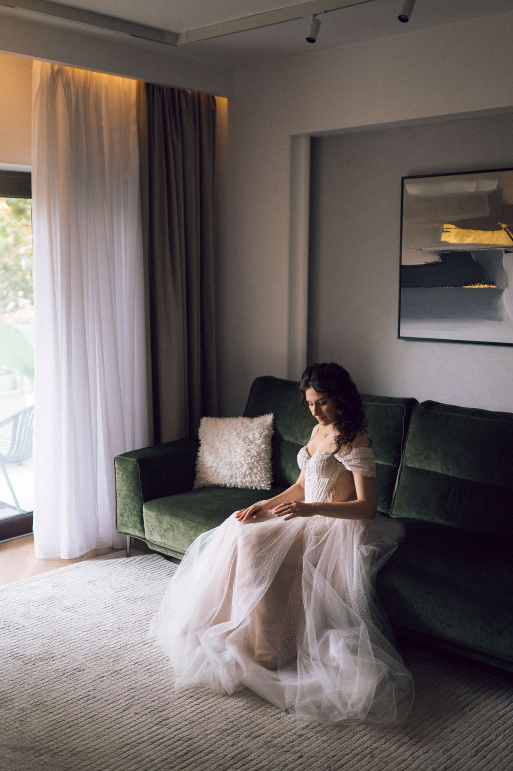 Bride in a flowing tulle wedding dress sitting gracefully on a green velvet sofa in an elegant hotel suite.