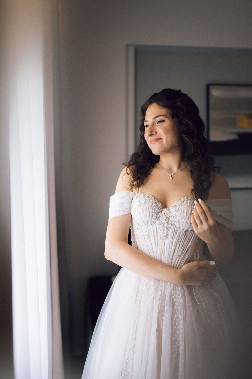 Smiling bride in off-shoulder lace wedding gown standing by window in soft natural light.