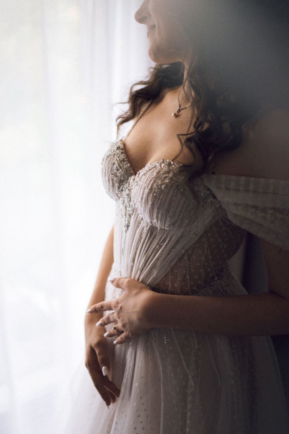 Bride in beaded off-shoulder wedding gown standing by window light, hands clasped at waist.