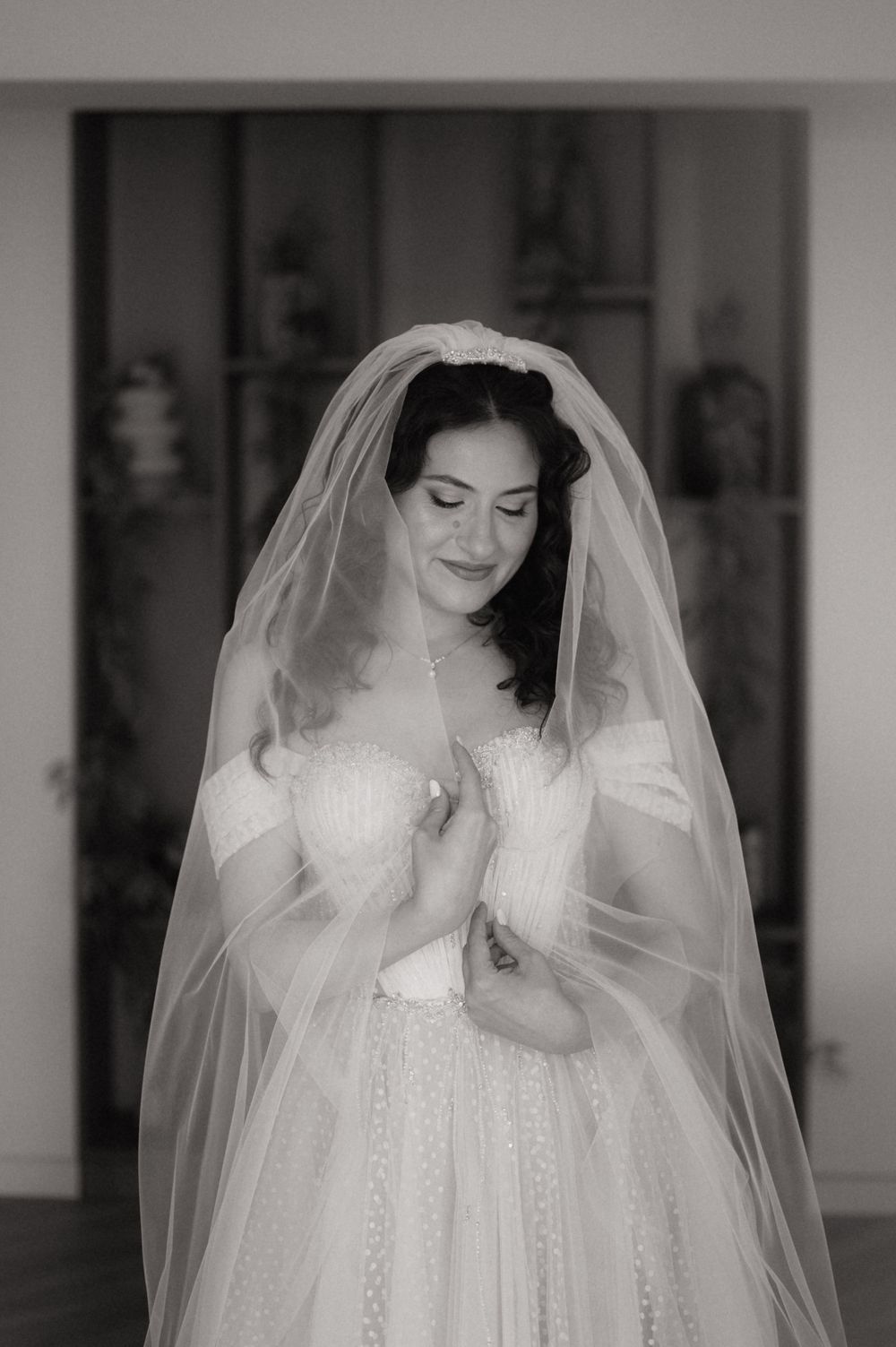 Black and white photo of a smiling bride in an off-shoulder gown and long veil, looking down elegantly.