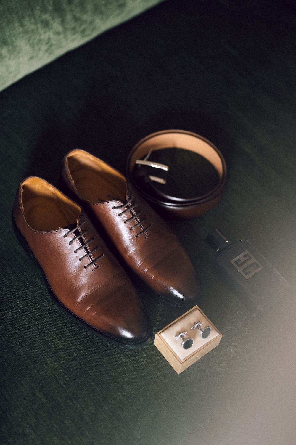 Brown leather Oxford dress shoes displayed with a dark leather belt and silver cufflinks in a wooden box on a green surface.