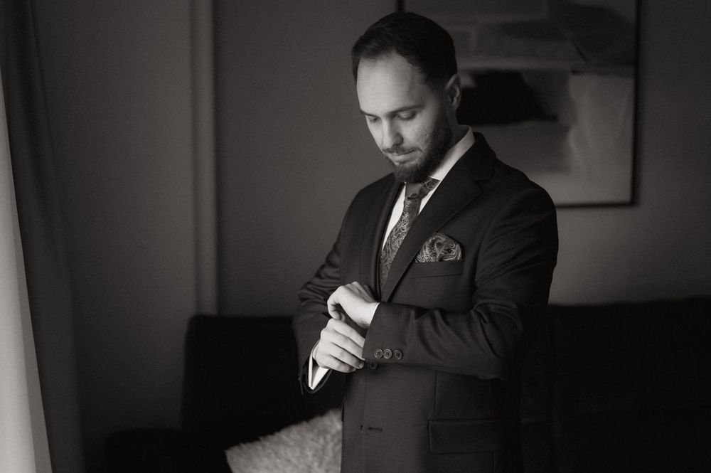 Elegant man in dark suit adjusting cufflinks in black and white photo, perfect for formal wedding attire inspiration.