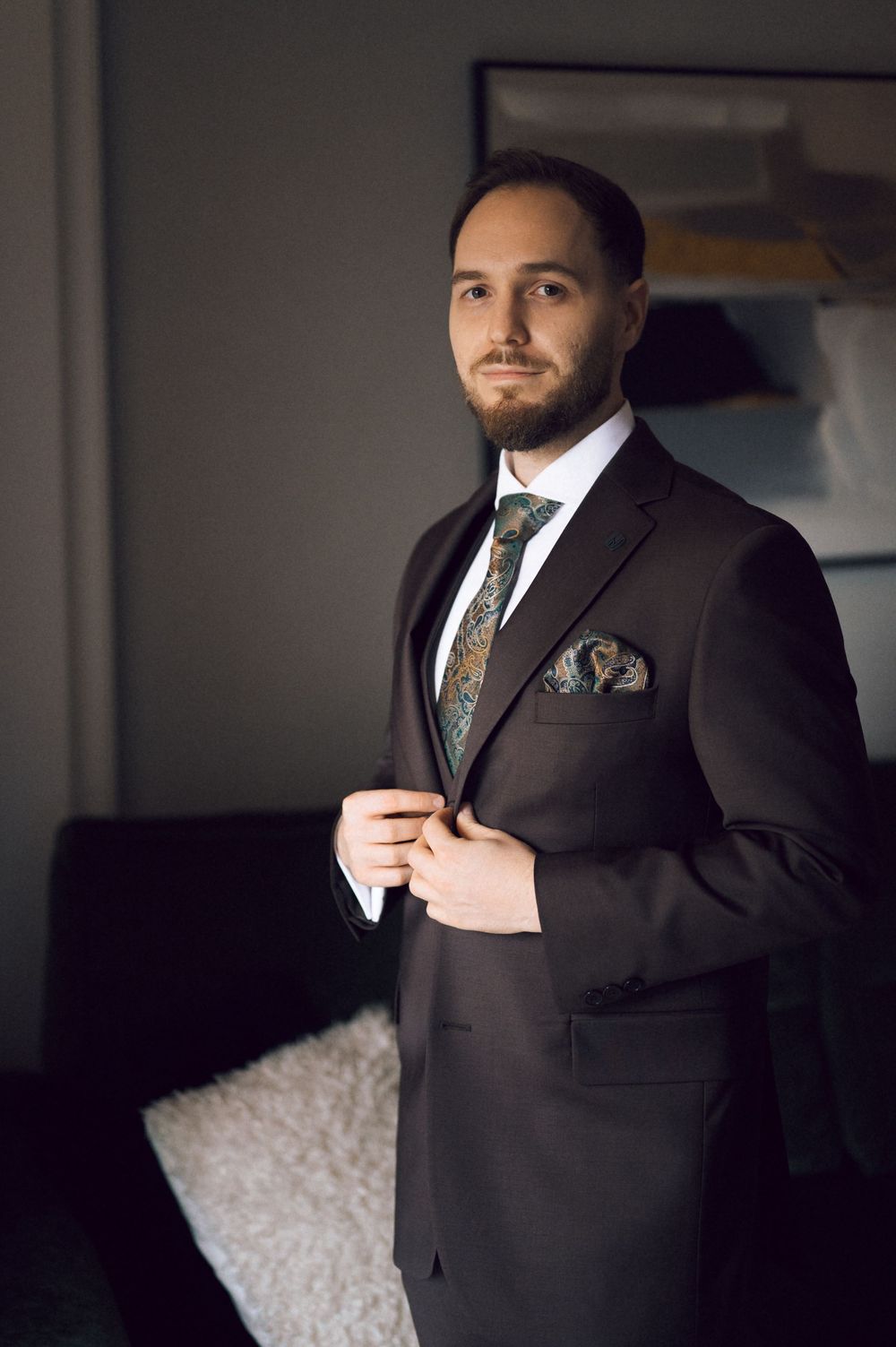 Man in dark suit adjusting jacket button, wearing paisley tie and matching pocket square.