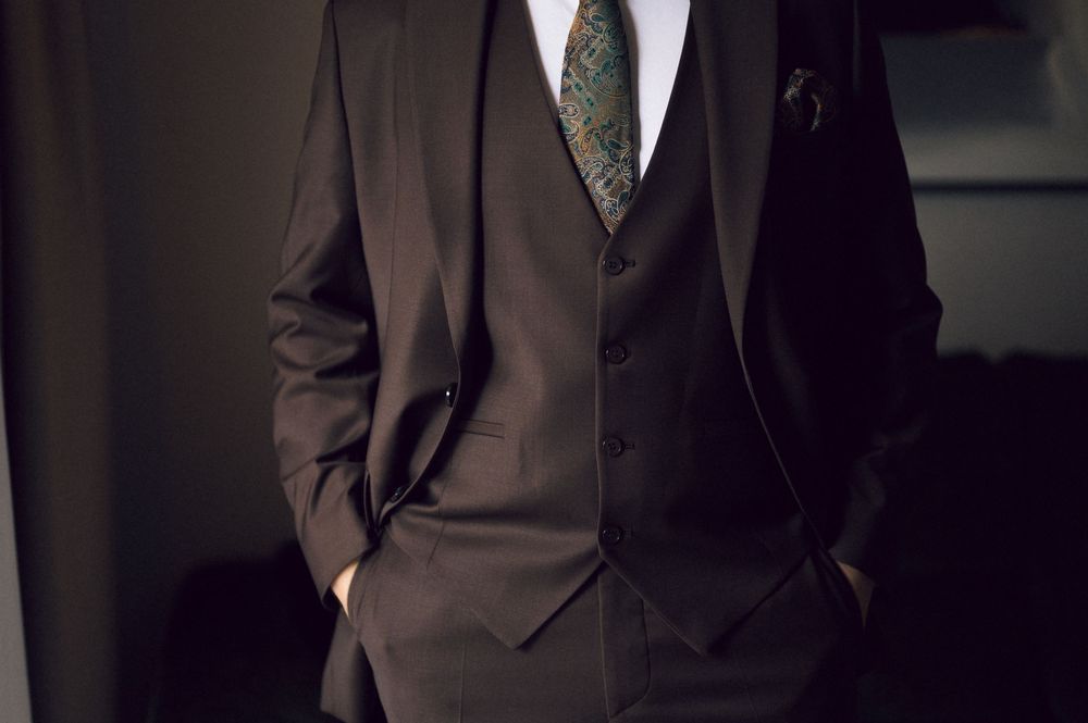 Man in dark brown three-piece suit with green tie and white dress shirt, hands in pockets, formal attire.