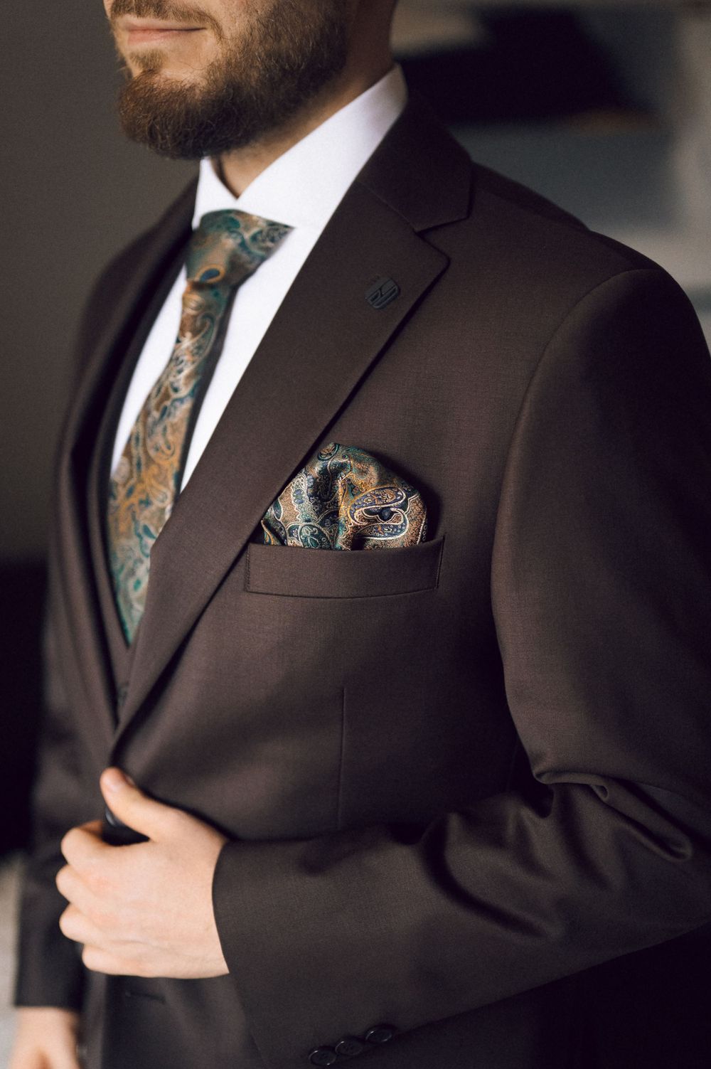 Close-up of dark suit with teal paisley tie and matching pocket square on bearded man.