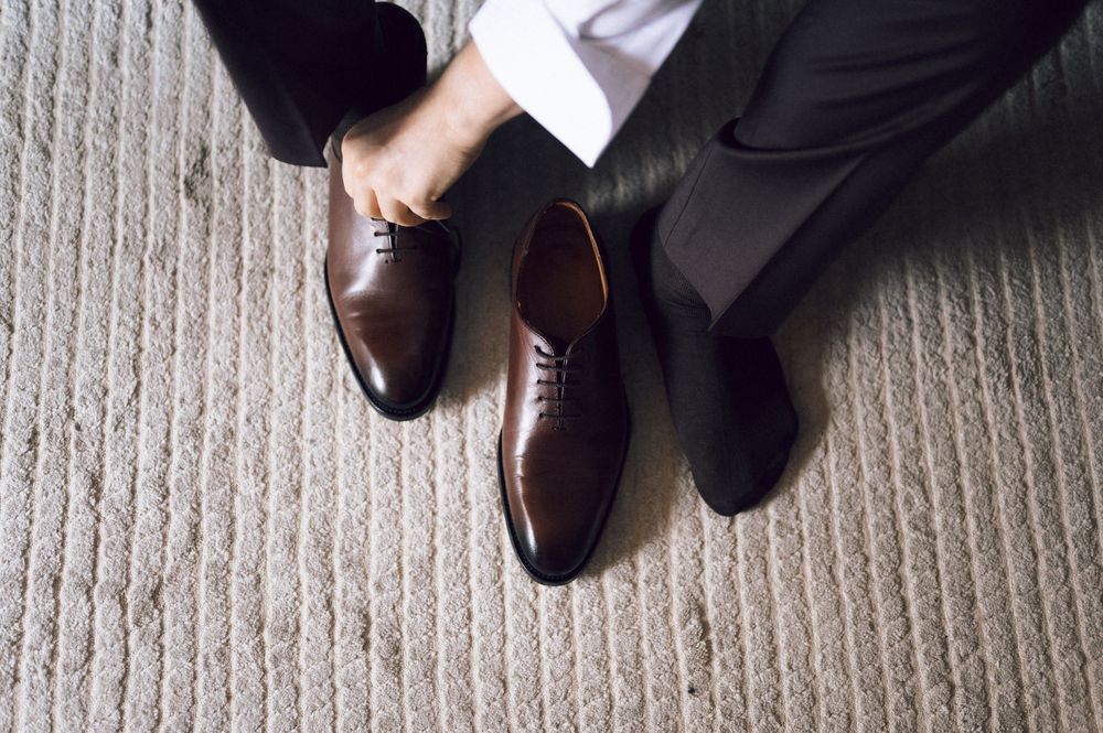 Man in dress pants putting on brown leather Oxford dress shoes on a textured carpet floor.